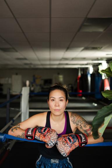 Stacy Leung stands in a boxing ring with her two arms up on the ropes with boxing globes on.