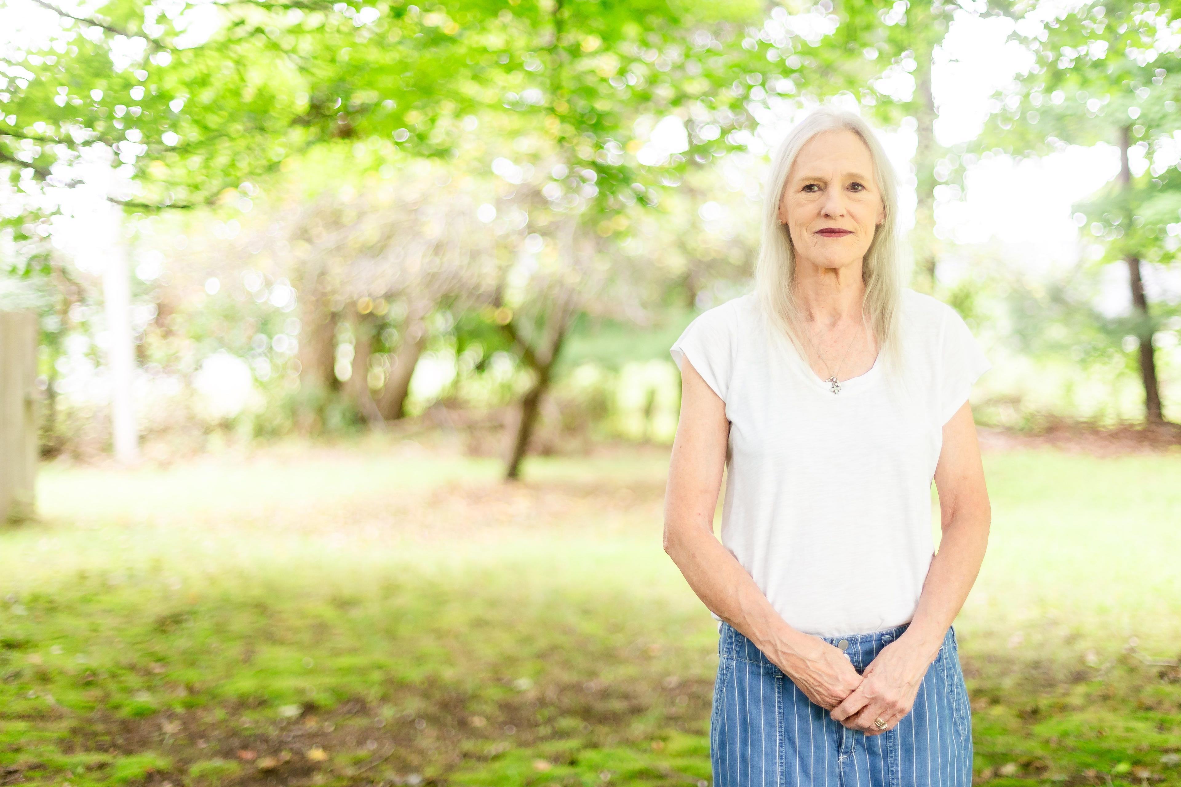 Jennifer Sanborn stands in front of trees and grass with a white T-shirt and blue jeans on.