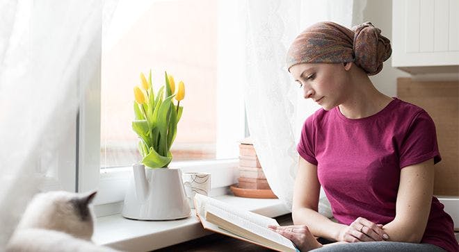 Image of a woman with a magenta t-shirt reading at a desk in her bedroom with the windows open.
